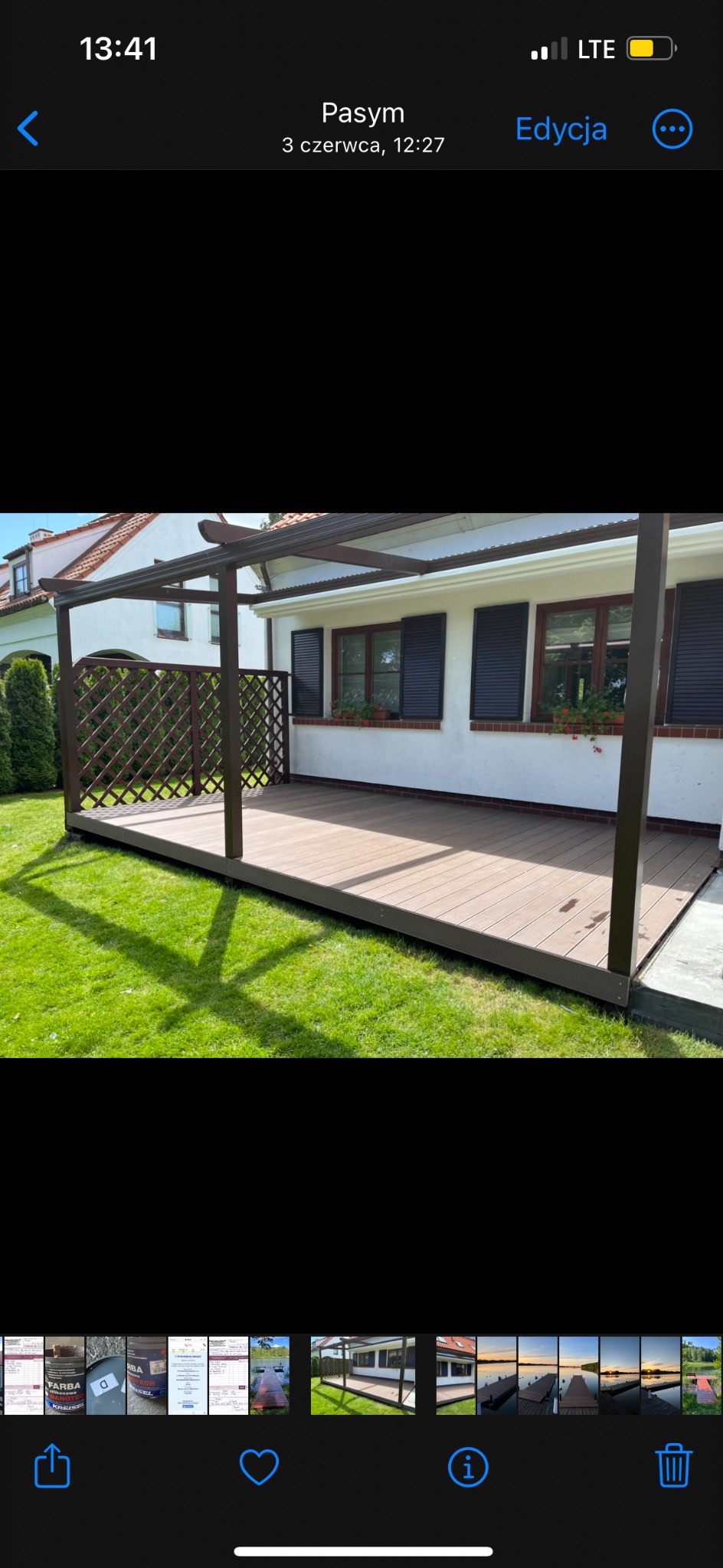 Zadaszony taras kompozytowy z brązową konstrukcją i ażurowym panelem, przylegający do białego domu z ciemnymi okiennicami, otoczony zielonym trawnikiem w słoneczny dzień.