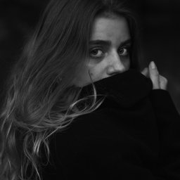 Vericones - Portret kobiety w czerni, z długimi włosami, ukrywającej usta za kołnierzem, w czarno-białej tonacji.