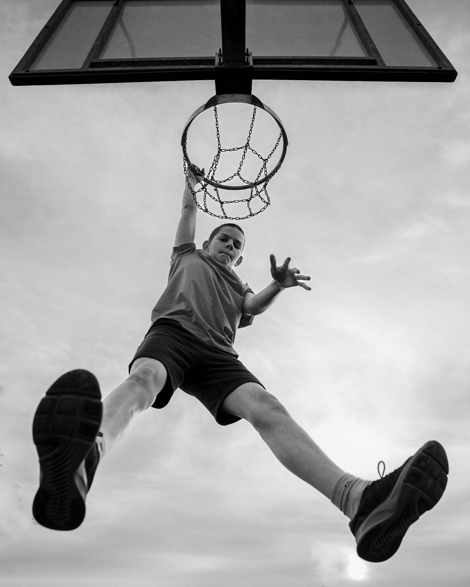 Czarno-białe ujęcie z dołu: nastolatek w locie, chwytający jedną ręką metalową siatkę kosza, druga ręka wyciągnięta, stopy z butami sportowymi skierowane w stronę obiektywu na tle pochmurnego nieba.
