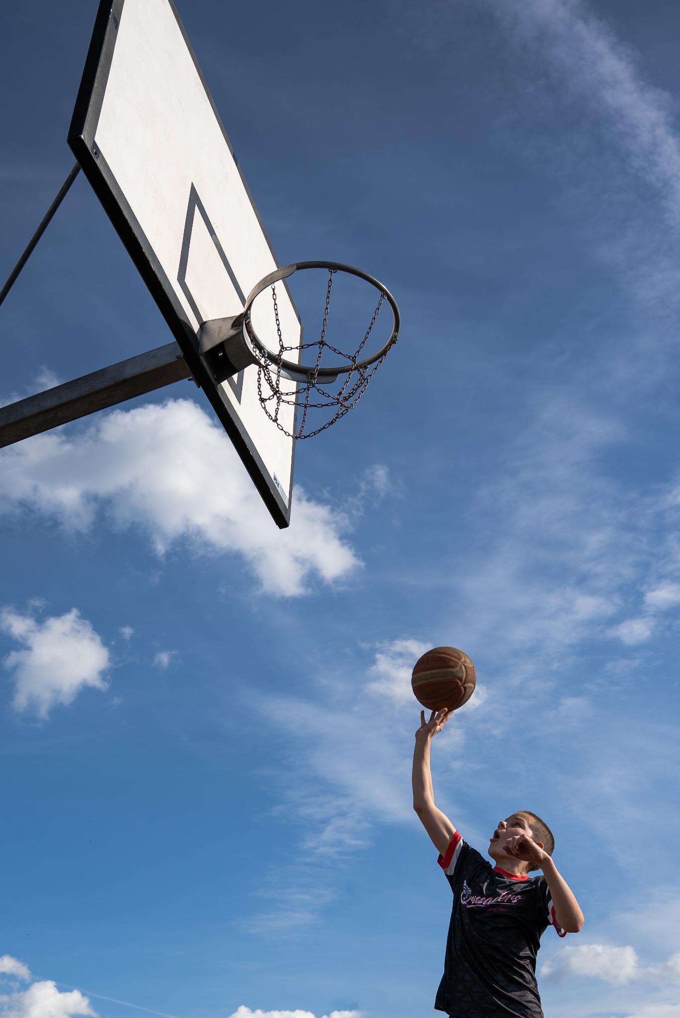 Młody koszykarz w czarnej koszulce z czerwonymi wstawkami oddaje rzut do kosza na tle błękitnego nieba z chmurami.