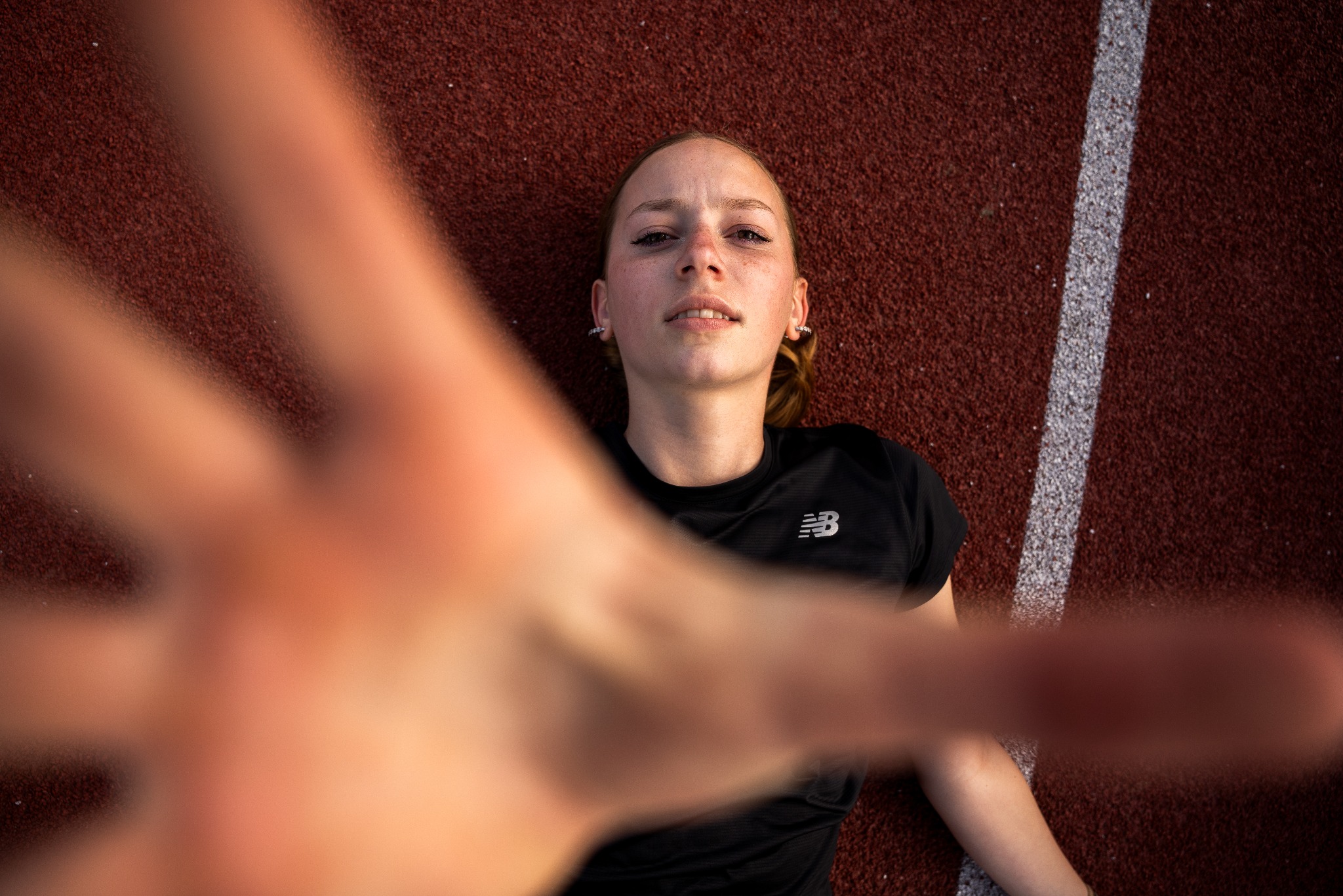 Młoda kobieta leżąca na bieżni tartanowej, ubrana w czarną koszulkę sportową z logo New Balance, wyciąga rękę w stronę obiektywu, tworząc efekt pierwszoosobowy.