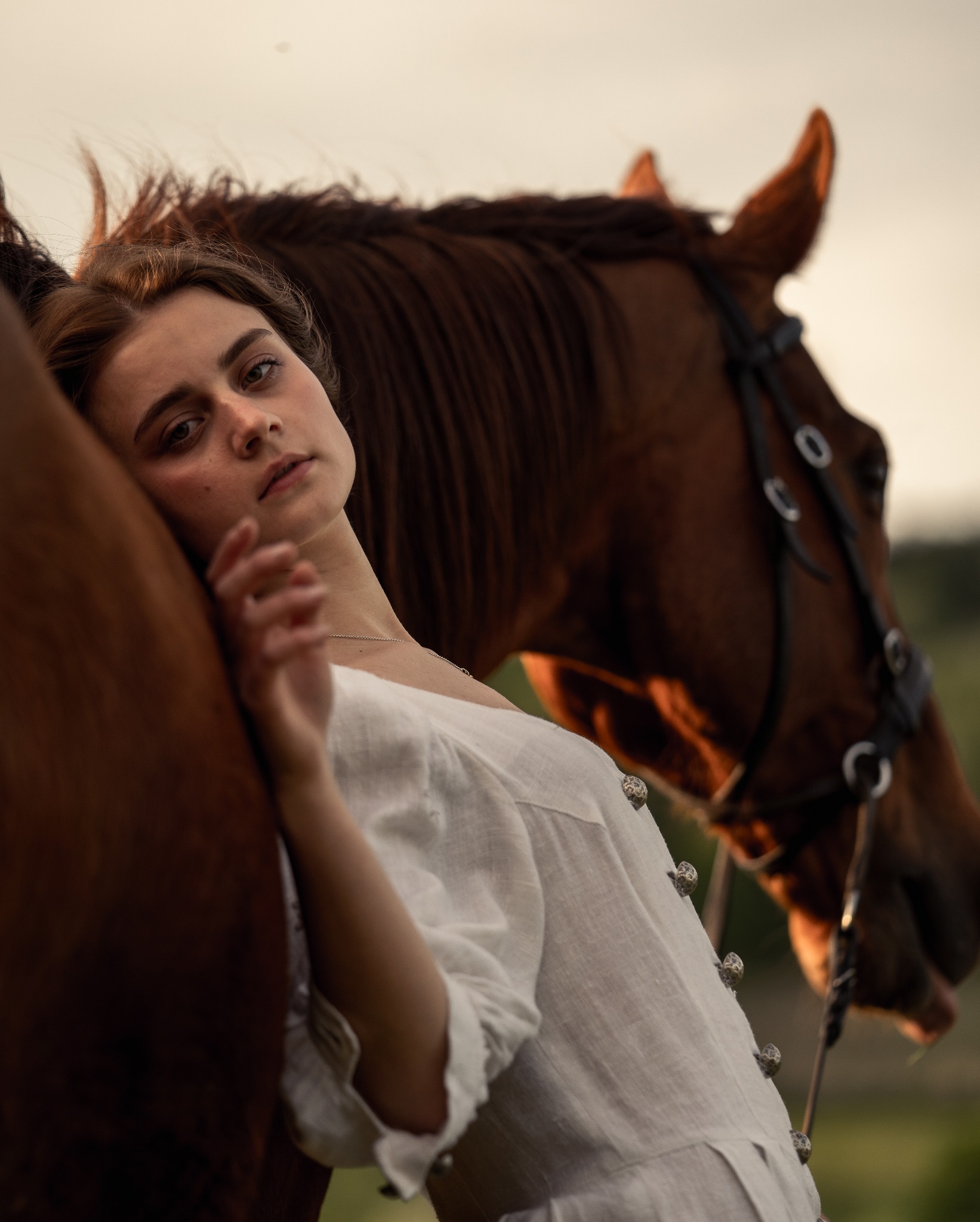 Portret kobiety w lnianej sukience opierającej się o brązowego konia z uprzężą na tle zieleni.