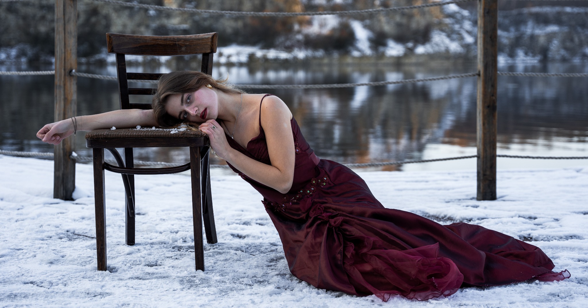 Kobieta w bordowej sukni wieczorowej opiera się o krzesło na śnieżnym pomoście z widokiem na zamarznięte jezioro w tle.