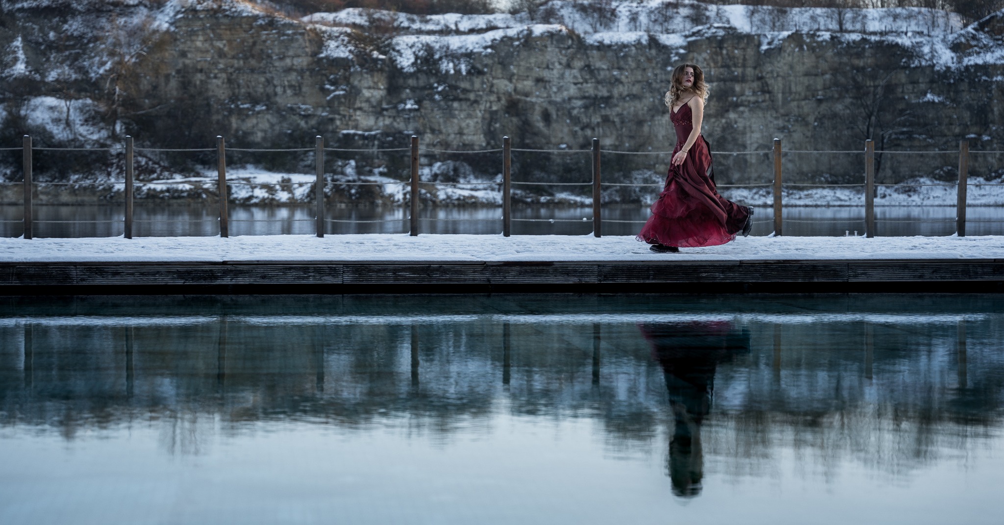 Kobieta w bordowej sukni spaceruje po drewnianym pomoście pokrytym śniegiem, z odbiciem w wodzie i skalistym tłem.