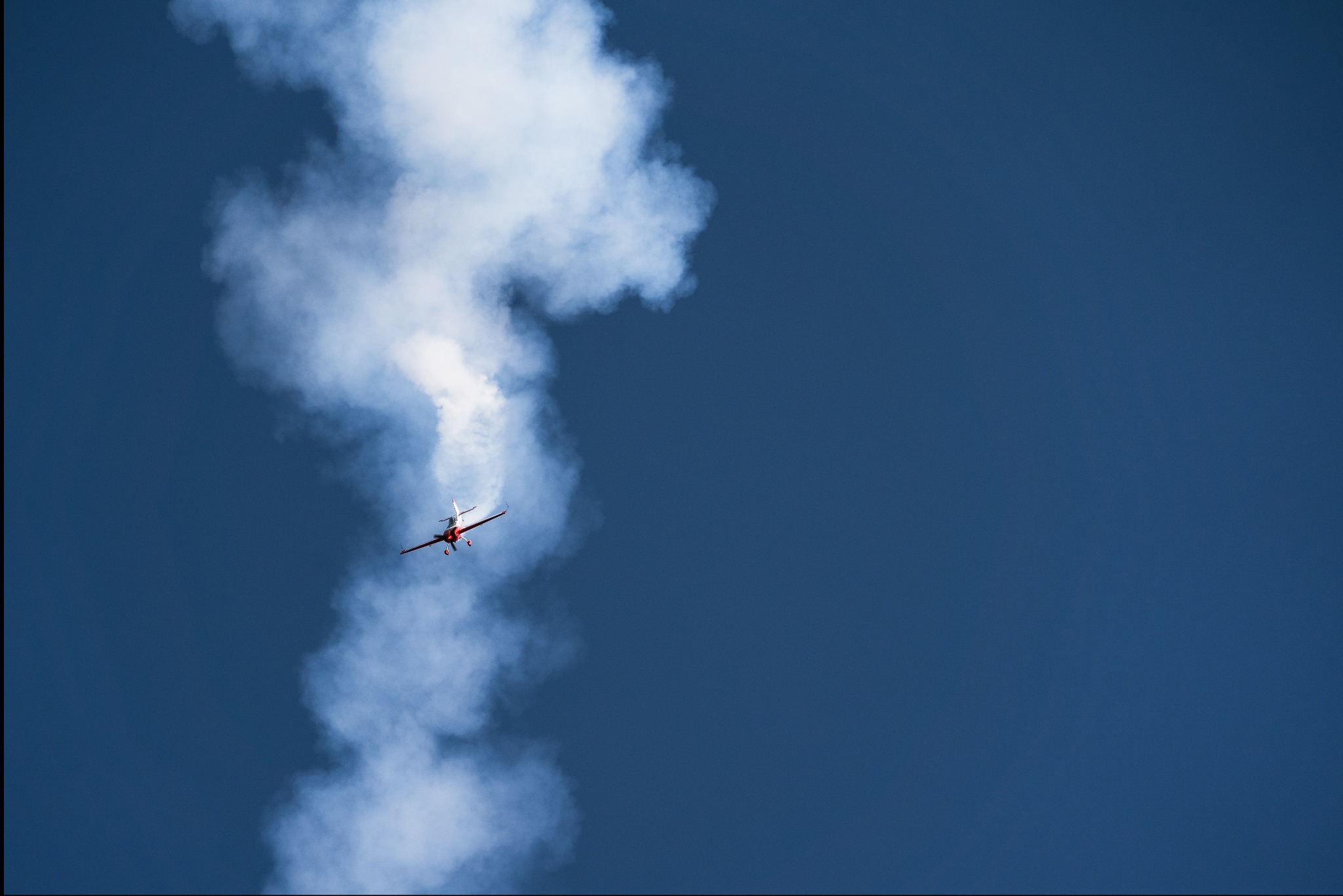 Czerwono-biały samolot akrobacyjny w locie na tle błękitnego nieba, pozostawiający za sobą ślad białego dymu.