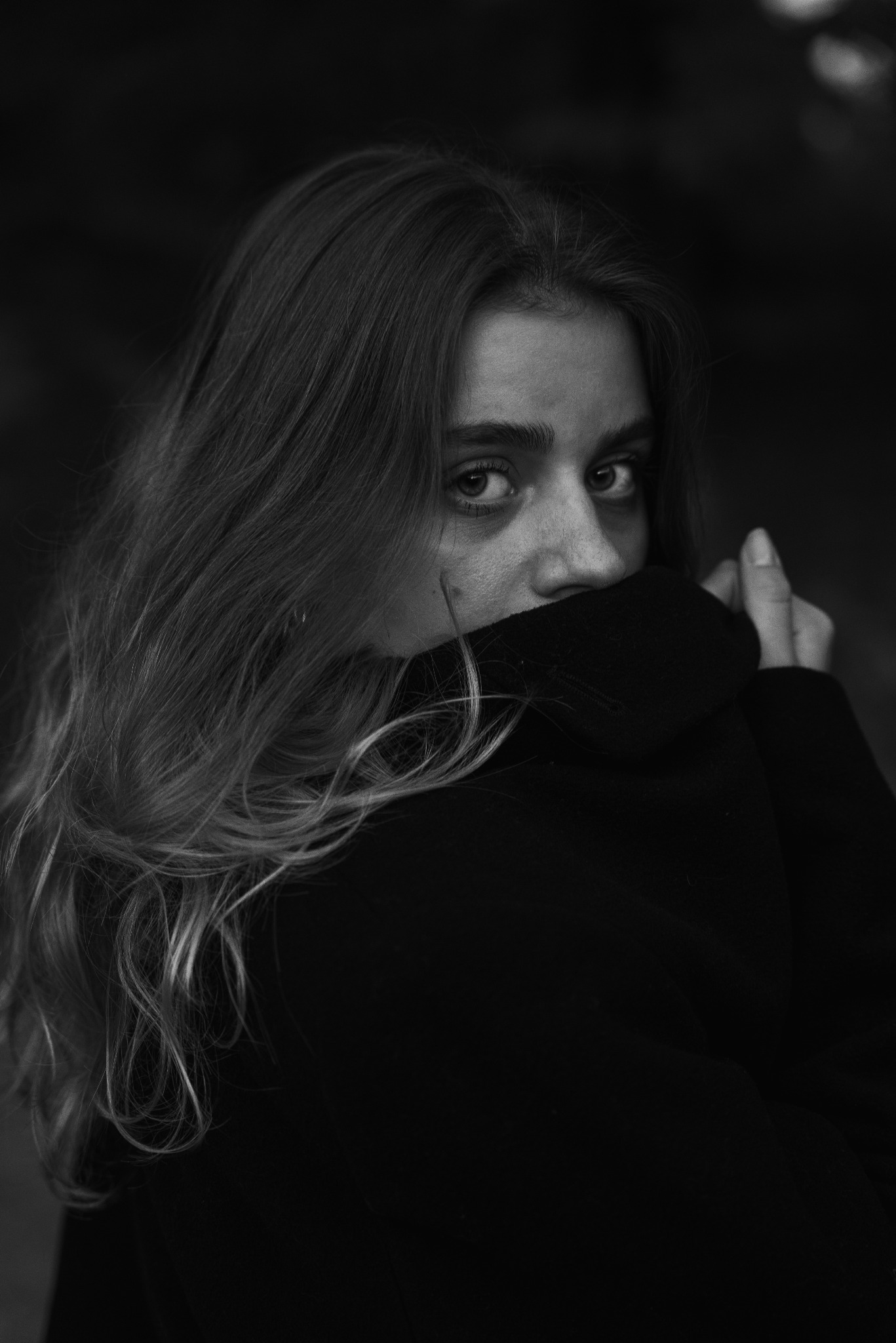 Portret kobiety w czerni, z długimi włosami, ukrywającej usta za kołnierzem, w czarno-białej tonacji.