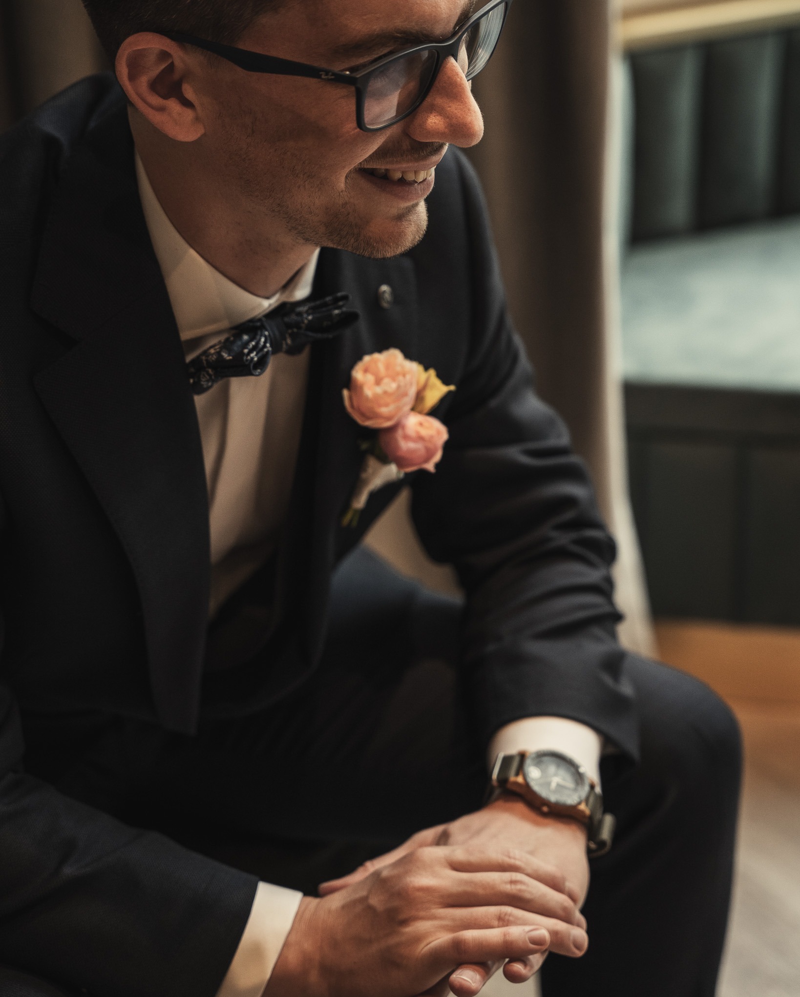 Uśmiechnięty mężczyzna w czarnym garniturze, z muszką i butonierką z różowych kwiatów, siedzący z założonymi rękami, na których widać zegarek z drewnianą bransoletą.