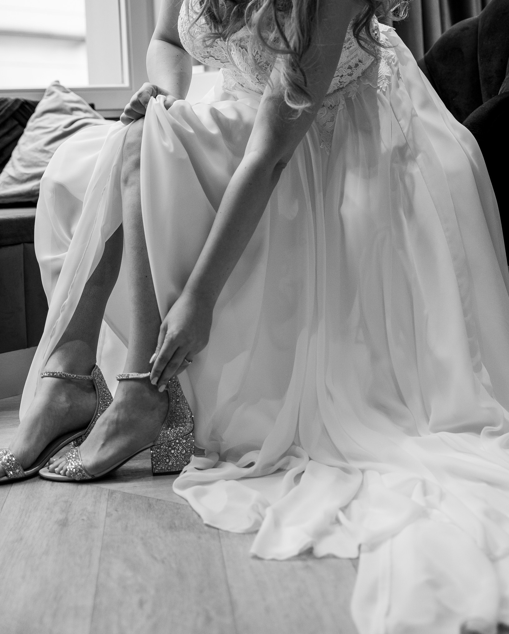 Panna młoda w białej sukni zapinająca brokatowe buty na obcasie, widoczne detale koronkowej góry sukni i pierścionek zaręczynowy.