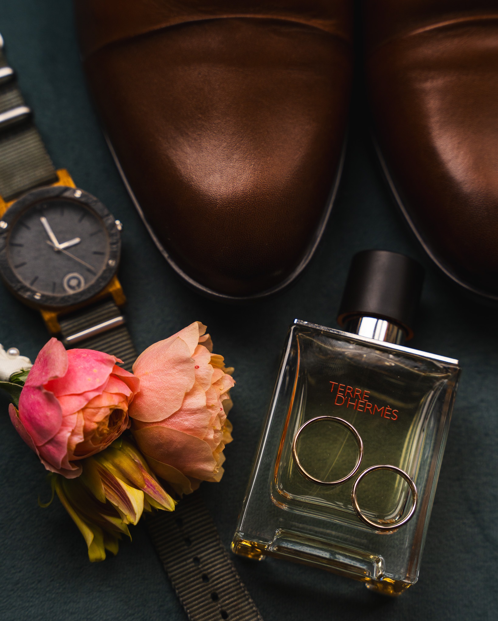Brązowe skórzane buty męskie, zegarek z paskiem w paski, butonierka z różowych i żółtych róż oraz flakon perfum Terre d'Hermès z dwoma złotymi obrączkami na tle ciemnego materiału.