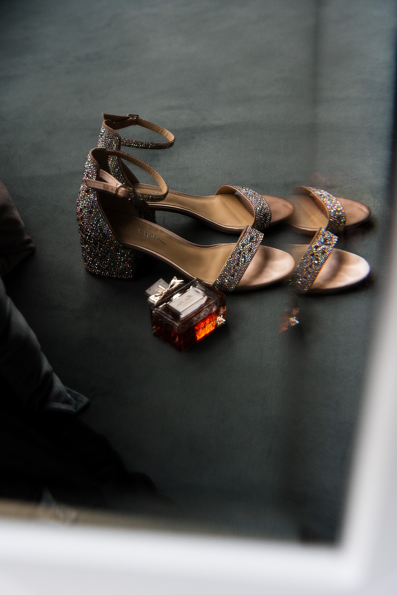 Para damskich, brokatowych sandałów na obcasie leżących obok flakonika perfum na ciemnym tle.