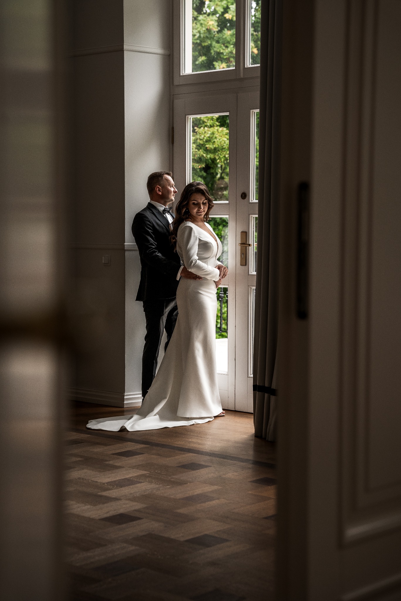 Elegancka para młoda w formalnych strojach, on w czarnym smokingu z muszką, ona w długiej białej sukni z rękawami, stojąca przy przeszklonych drzwiach z widokiem na zieleń, z delikatnym...