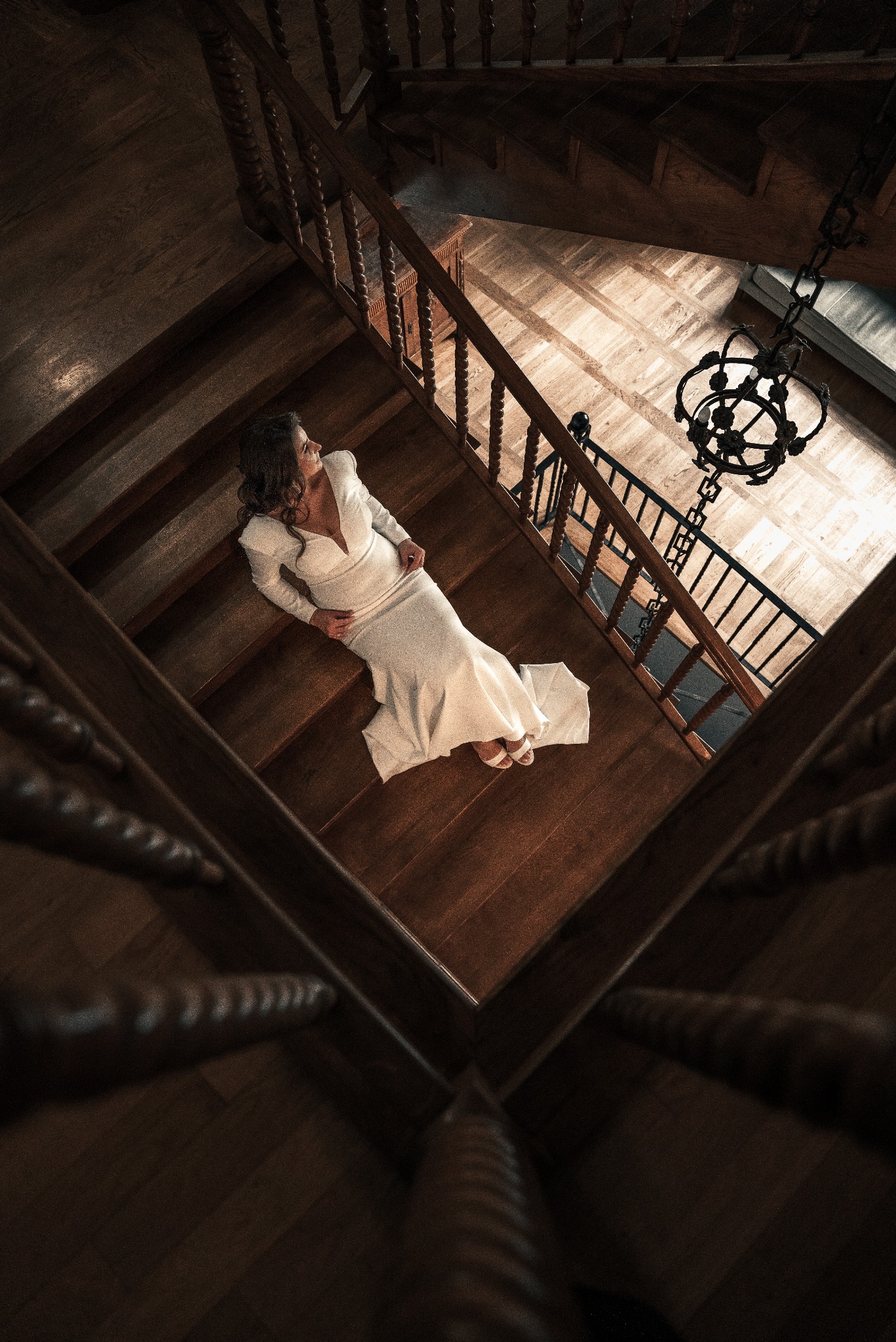 Kobieta w białej sukni leży na drewnianych schodach, widok z góry na spiralne schody i żyrandol