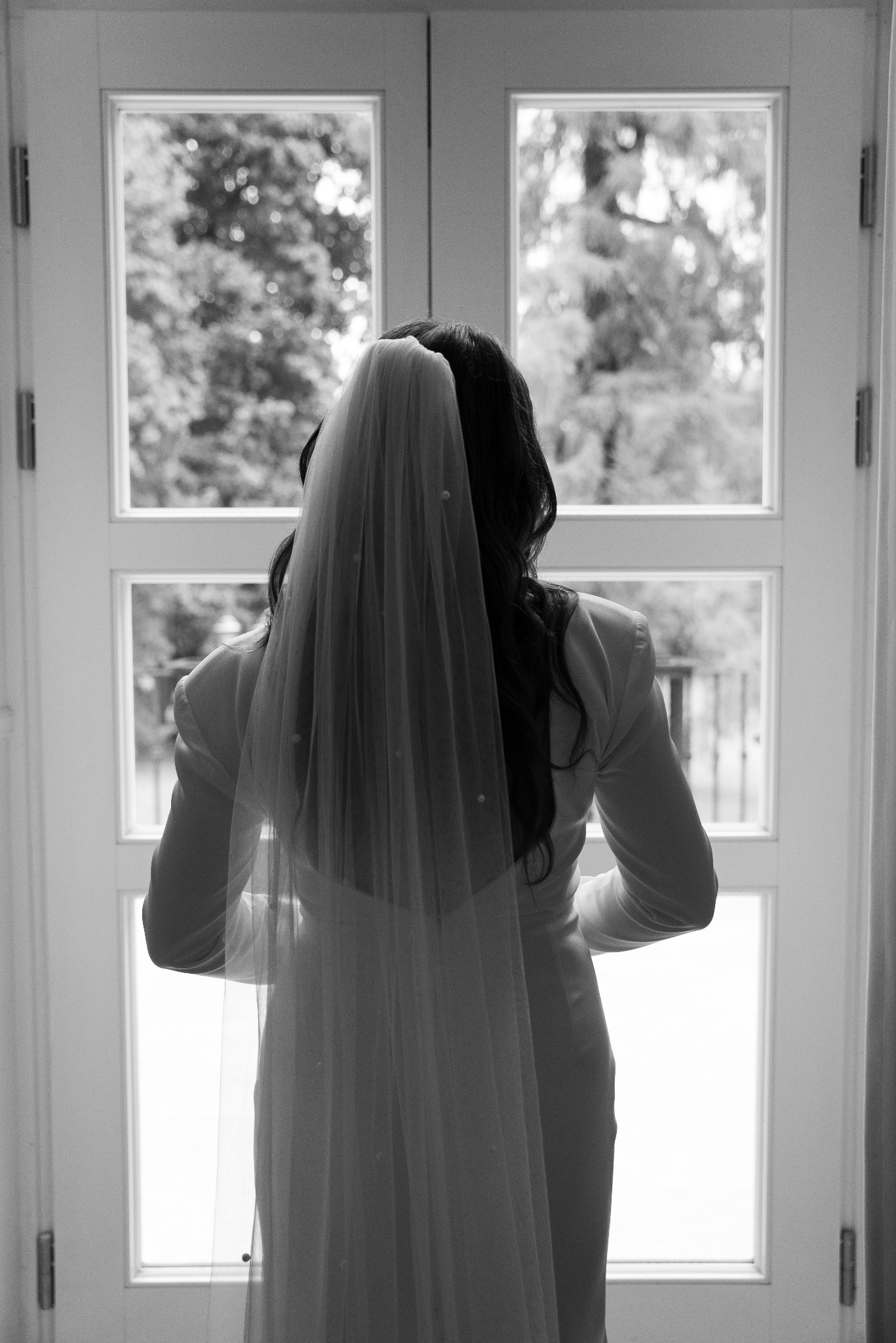 Panna młoda w białym garniturze i welonie, stojąca tyłem do kamery przed przeszklonymi drzwiami z widokiem na zieleń, czarno-biała fotografia.