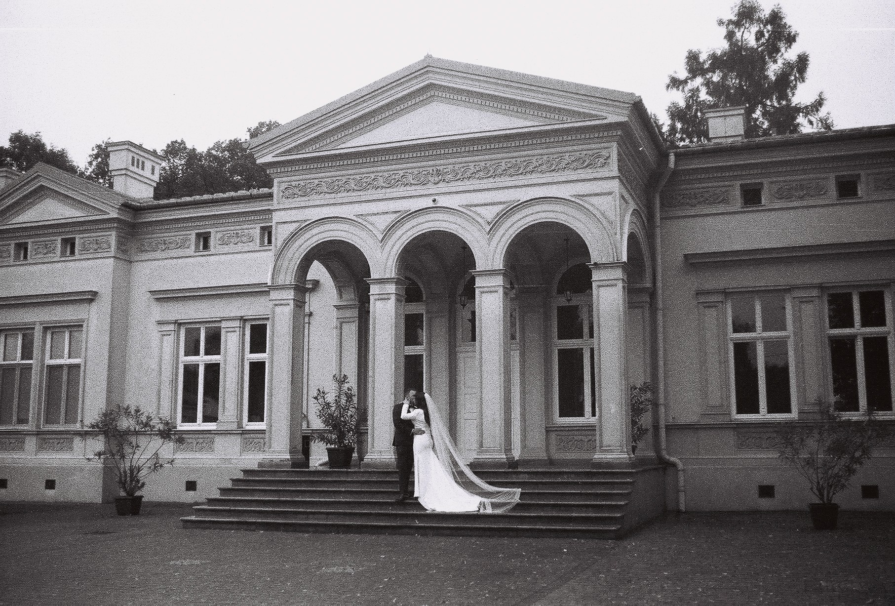 Czarno-białe zdjęcie pary młodej całującej się na schodach klasycystycznego budynku z kolumnami i łukami, długa woalka panny młodej opada na schody.