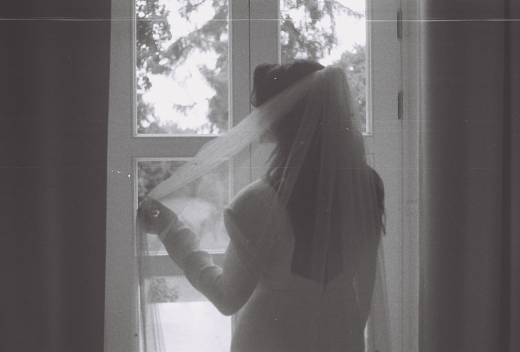 Czarno-białe zdjęcie panny młodej w sukni ślubnej i welonie, stojącej tyłem do kamery przy oknie z widokiem na zieleń.