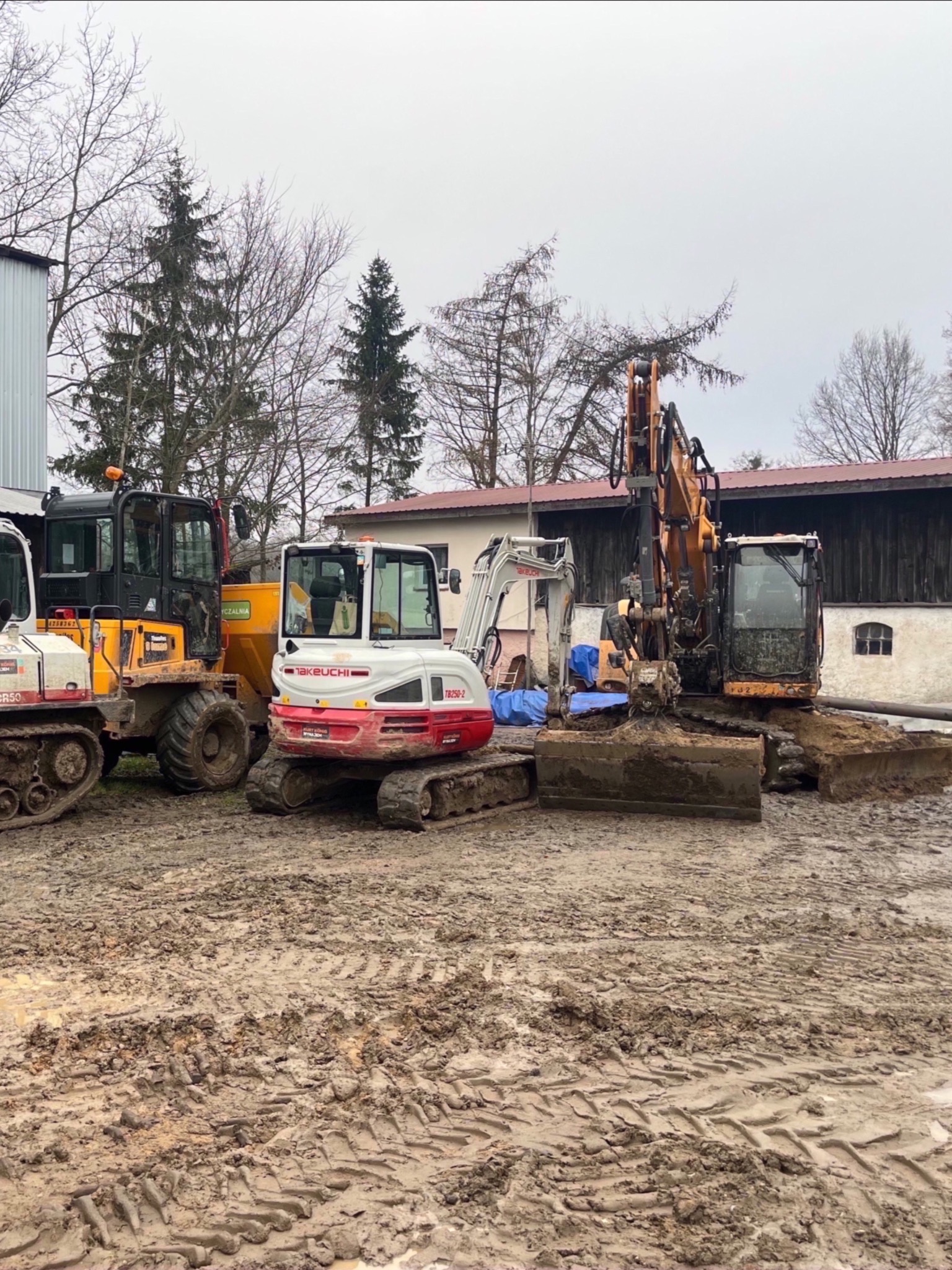 Park maszyn budowlanych: koparki i ładowarki w błotnistym terenie. Takeuchi TB250-2 na pierwszym planie, inne maszyny w tle. Szare niebo, budynek w tle.