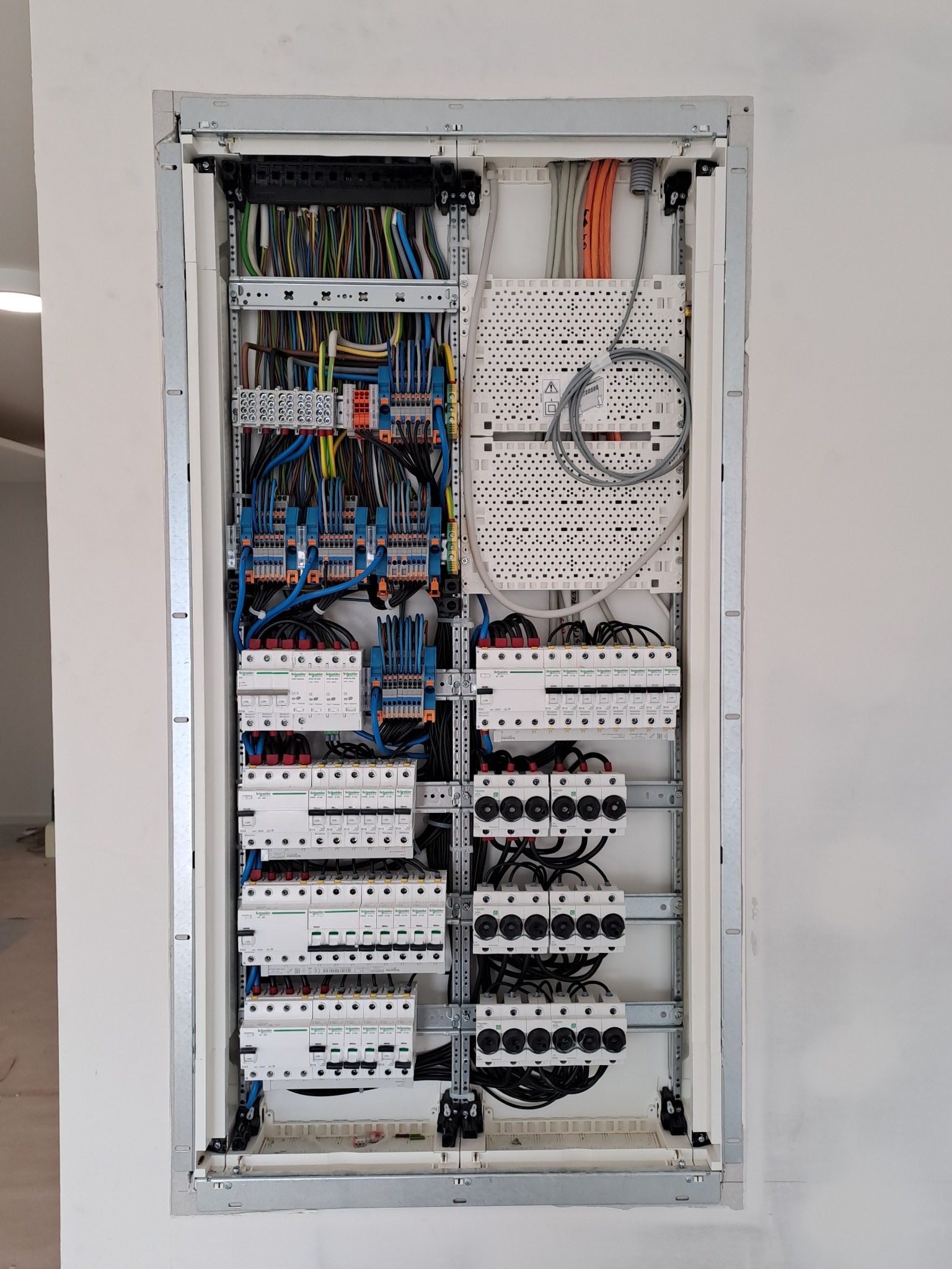 Otwarta skrzynka rozdzielcza z widocznymi bezpiecznikami, przewodami w różnych kolorach oraz elementami montażowymi, zamontowana w ścianie w surowym stanie.