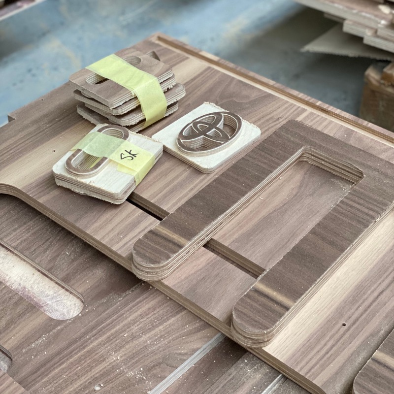 Wycięte z drewna elementy, w tym ramki i logo motoryzacyjne, ułożone na drewnianej płycie z widocznymi śladami obróbki.