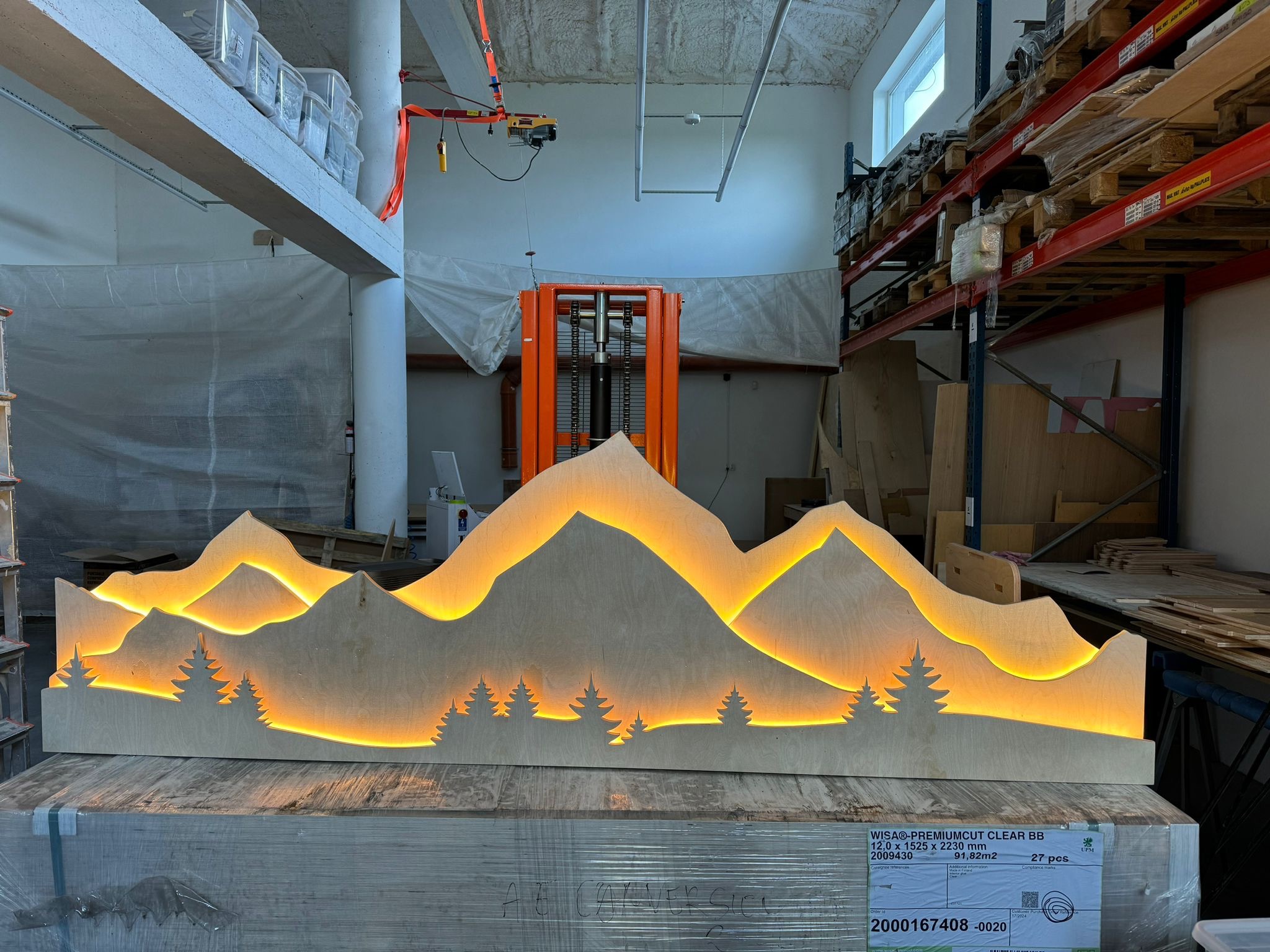 Wycięty z drewna, podświetlany motyw górski z sylwetkami drzew iglastych, stojący na palecie z widocznym tekstem 'WISA PREMIUMCUT CLEAR BB', w tle magazyn z wózkiem widłowym