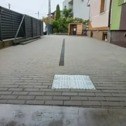 Szara kostka brukowa ułożona na podjeździe, z widoczną metalową kratką drenażową pośrodku i metalową studzienką rewizyjną, brama w tle, pochmurny dzień.