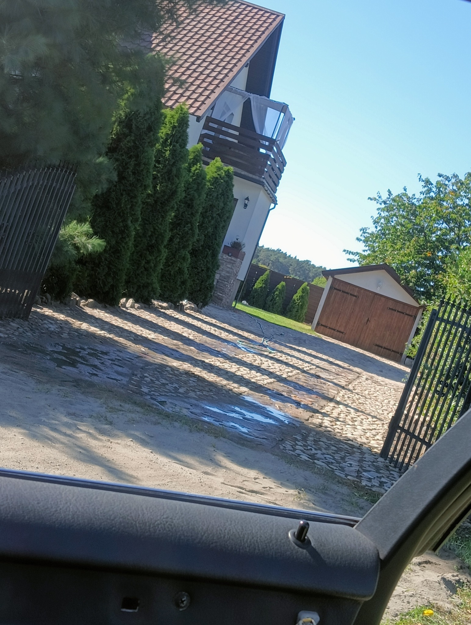 Widok z wnętrza samochodu na posesję z kostką brukową i domem w słoneczny dzień. Na kostce leżą cienie drzew, widoczne ślady po wodzie.