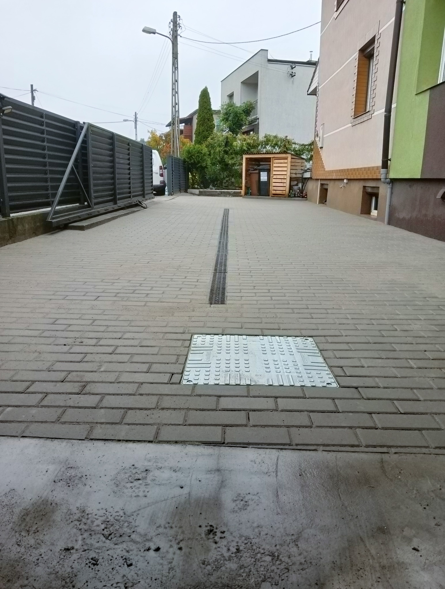 Szara kostka brukowa ułożona na podjeździe, z widoczną metalową kratką drenażową pośrodku i metalową studzienką rewizyjną, brama w tle, pochmurny dzień.