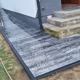 MegaR twój dom i ogród - Ułożona ścieżka z szarych płyt imitujących drewno, prowadząca do schodów wejściowych. Widoczny piasek i trawa, co sugeruje świeżo zakończone prace.