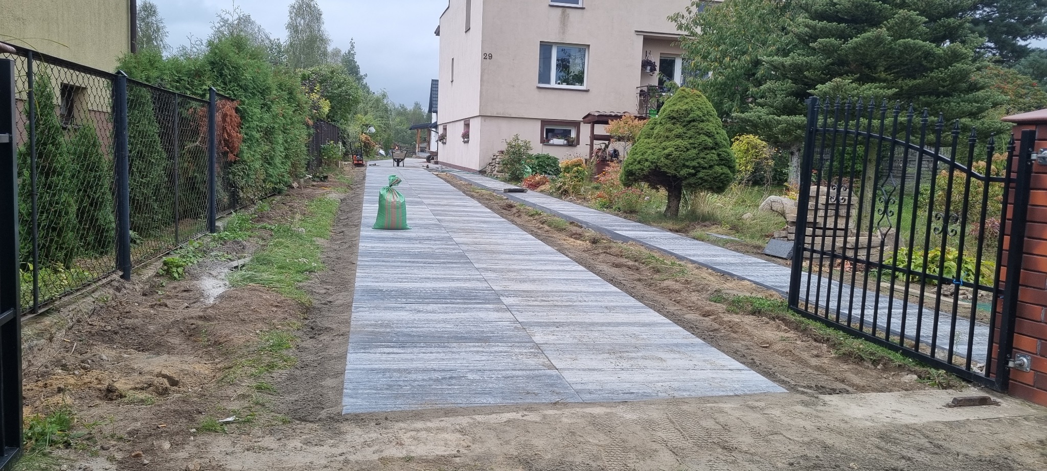 Ułożona kostka brukowa tworząca ścieżkę w ogrodzie, widoczne narzędzia i materiały budowlane, brama w tle, dom z numerem 29.