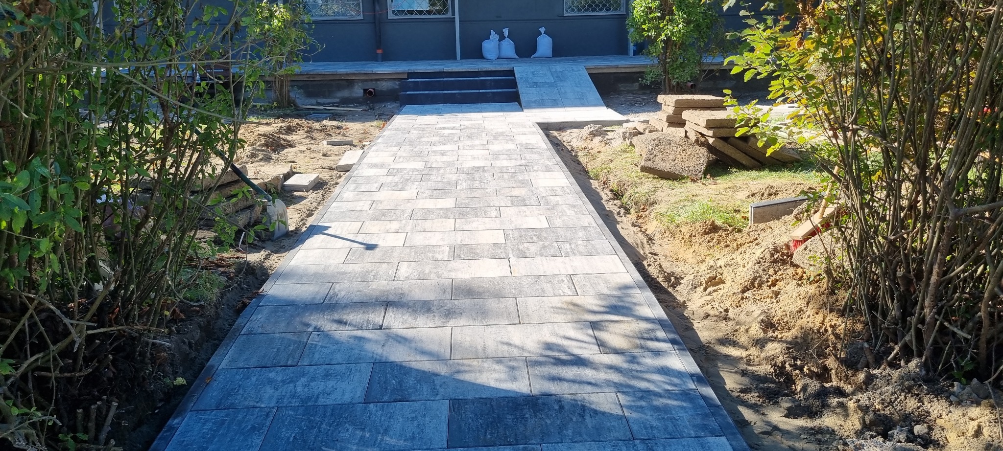 Ułożona ścieżka z szarej kostki brukowej prowadząca do domu z schodami. Częściowo widoczne rośliny i narzędzia. Słoneczny dzień.