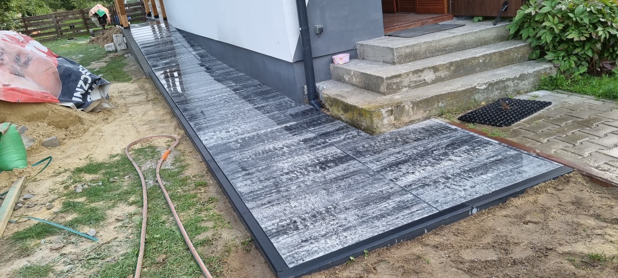Ułożona ścieżka z szarych płyt imitujących drewno, prowadząca do schodów wejściowych. Widoczny piasek i trawa, co sugeruje świeżo zakończone prace.