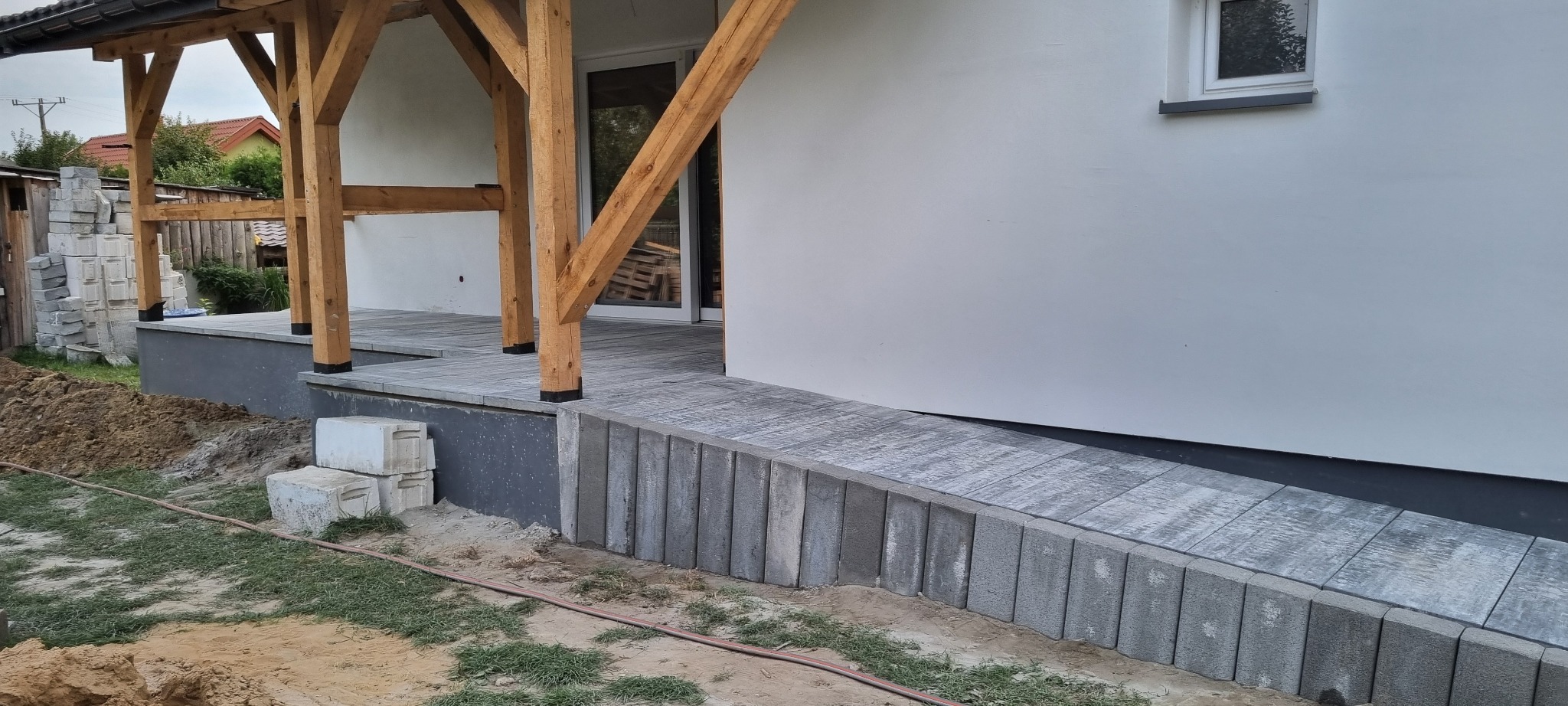 Wykończony taras z kostki brukowej przy domu z drewnianą konstrukcją zadaszenia. Widoczne elementy kostki, ściana budynku i częściowo trawnik.