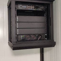 PE-INSTAL PAWEŁ JABŁOŃSKI - Czarna szafa rack Lanberg z zamontowanym switchem, listwą zasilającą i innymi urządzeniami, zainstalowana na ścianie z paneli. Widoczne kable i gniazda.