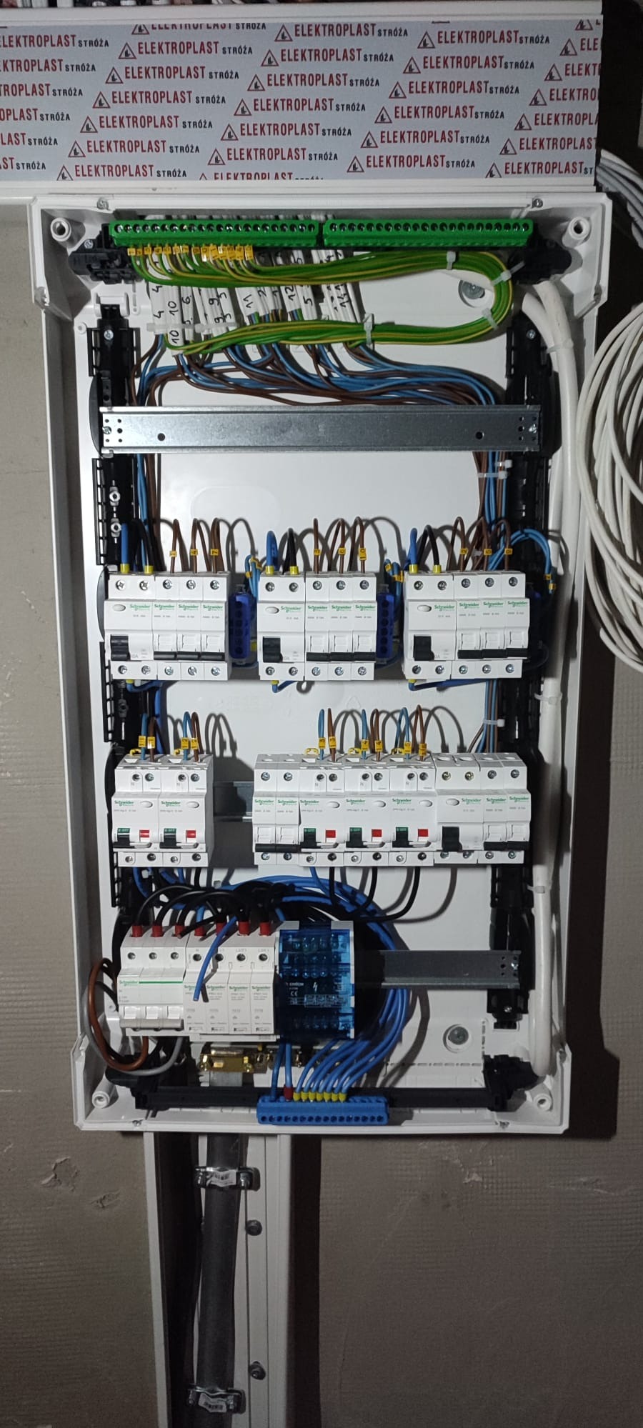 Rozdzielnica elektryczna z zainstalowanymi bezpiecznikami, przewodami i listwami zaciskowymi w białej obudowie. Widoczne elementy marki Elektroplast.