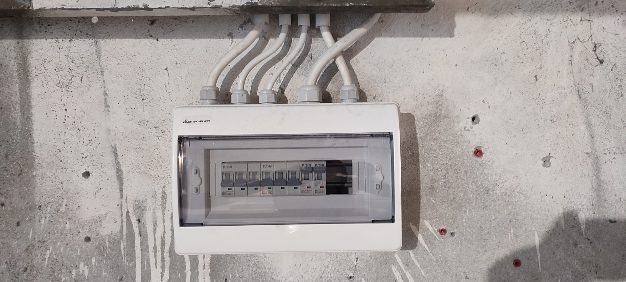 Zamontowana, biała skrzynka elektryczna Elektro-Plast z widocznymi bezpiecznikami Eaton, przymocowana do surowej, betonowej ściany z kablami w białych peszlach.