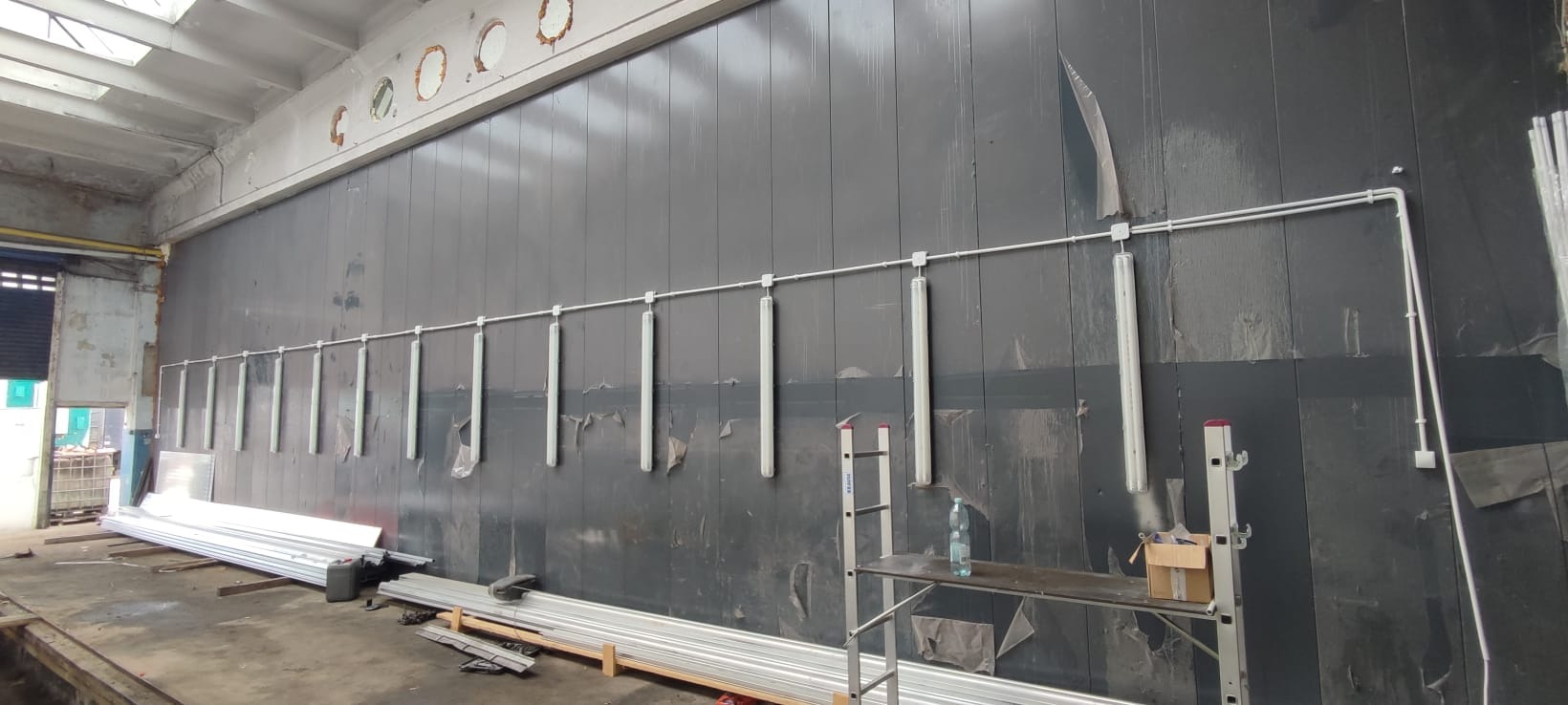 Długa ściana z zamontowanym rzędem lamp LED i instalacją elektryczną w rurkach. Widoczne rusztowanie i materiały budowlane, surowy stan wykończenia wnętrza.