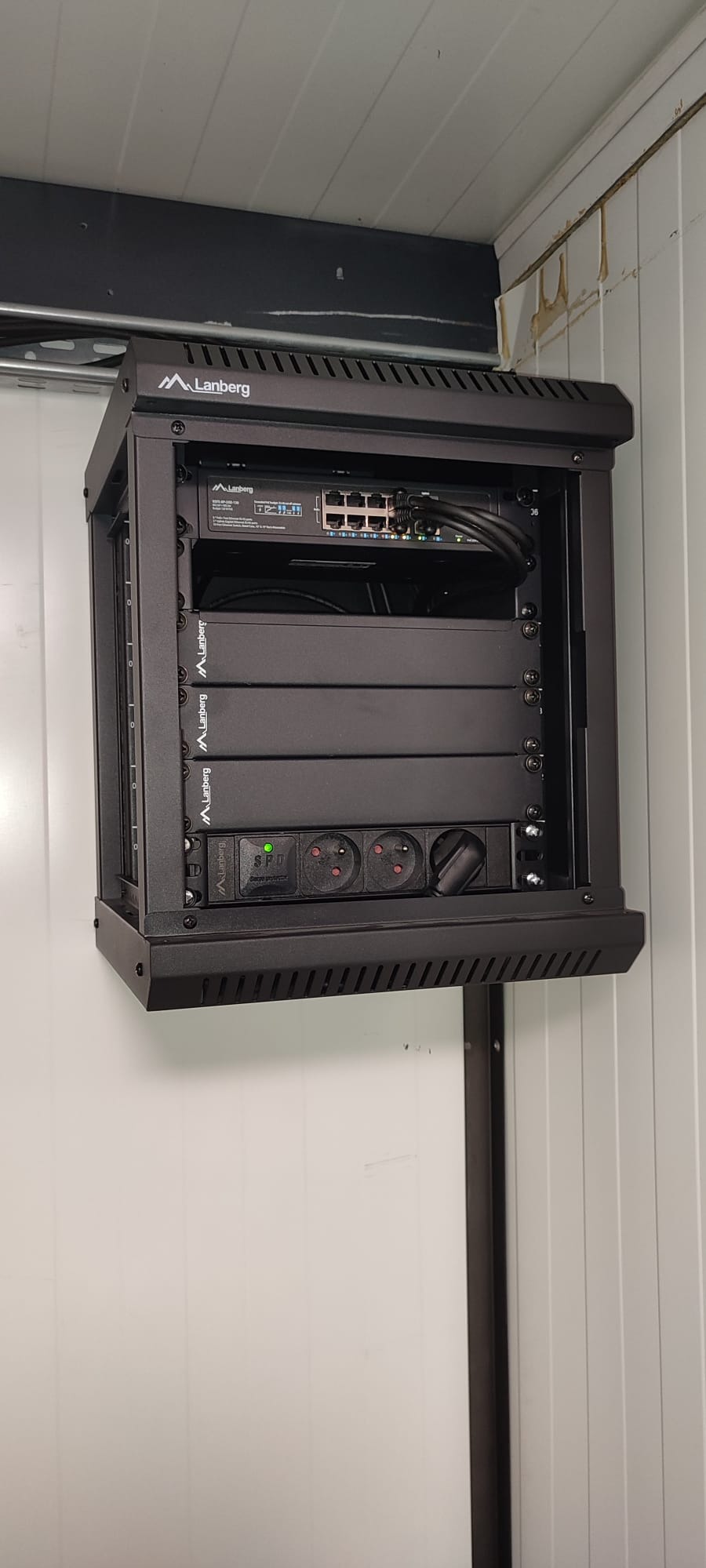 Czarna szafa rack Lanberg z zamontowanym switchem, listwą zasilającą i innymi urządzeniami, zainstalowana na ścianie z paneli. Widoczne kable i gniazda.