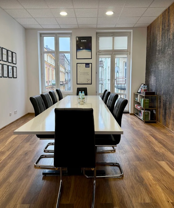 Sala konferencyjna z długim białym stołem, czarnymi krzesłami i widokiem na ulicę przez okna. Na ścianach wiszą oprawione dokumenty i plakaty.