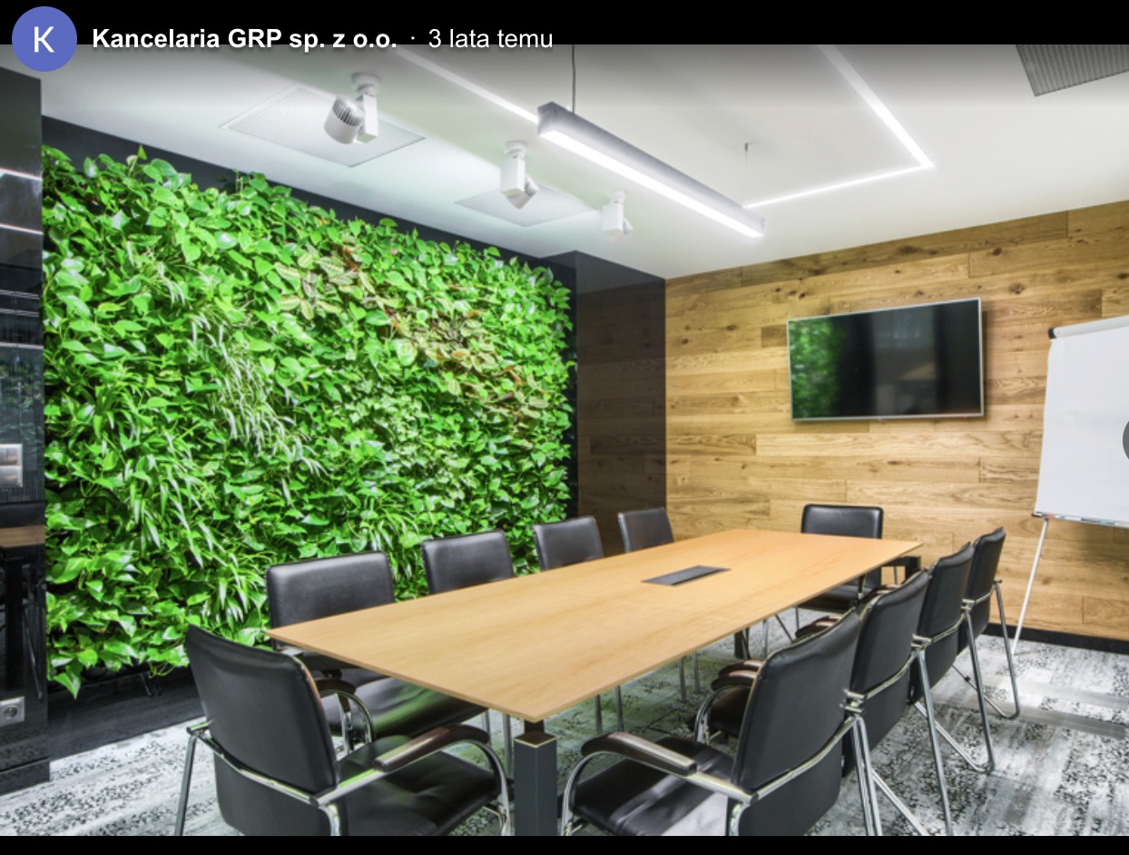 Sala konferencyjna z drewnianą ścianą, zieloną ścianą z roślin i długim stołem z czarnymi krzesłami. Nowoczesne oświetlenie sufitowe.
