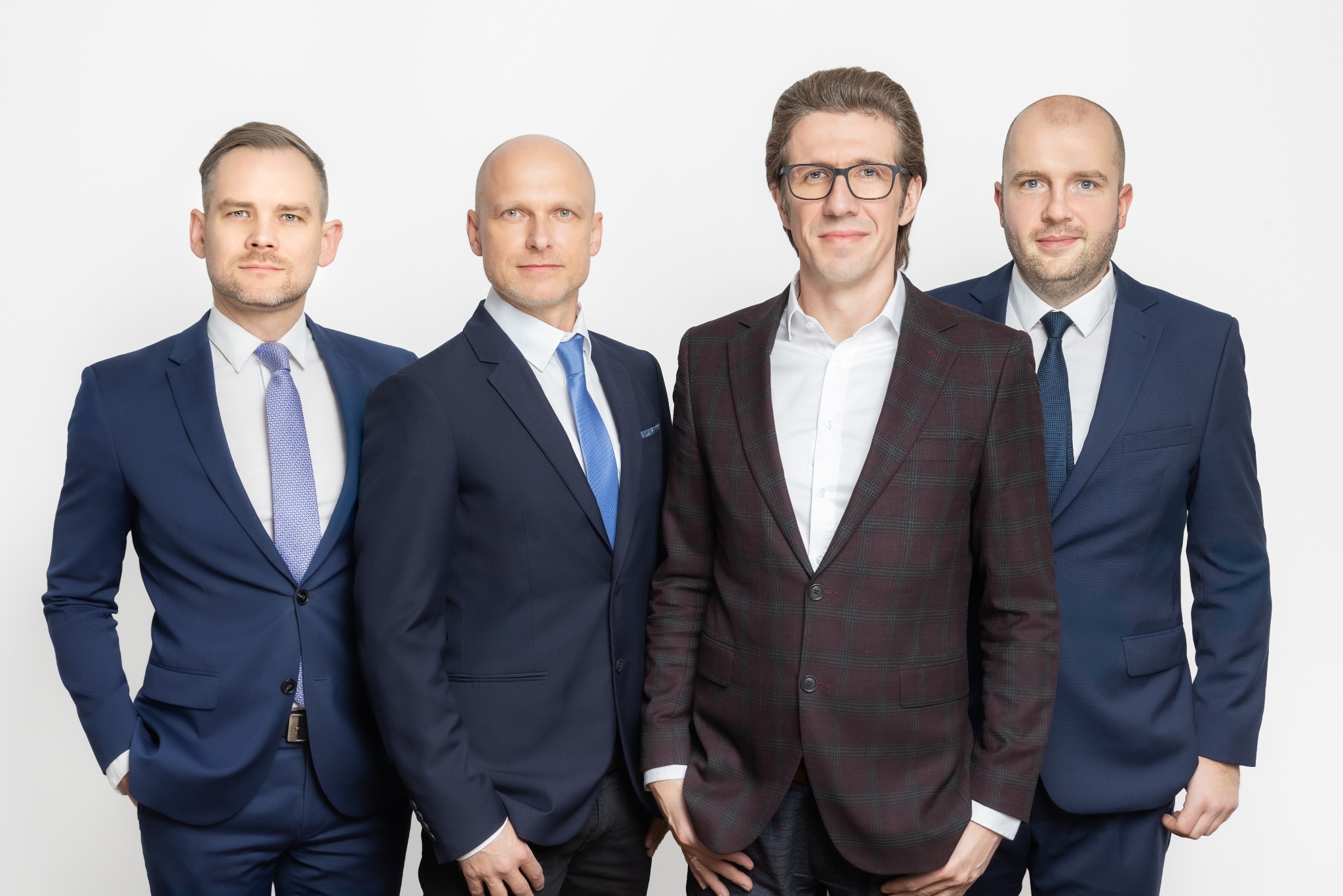 Portret grupowy czterech mężczyzn w garniturach na białym tle. Jeden z nich ma na sobie bordową marynarkę w kratę i okulary. Pozostali w niebieskich garniturach.