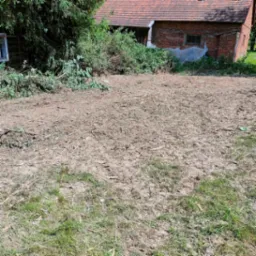 Teren po wycince drzew i krzewów, widoczne pozostałości gałęzi i pni na tle starego, ceglanego budynku.