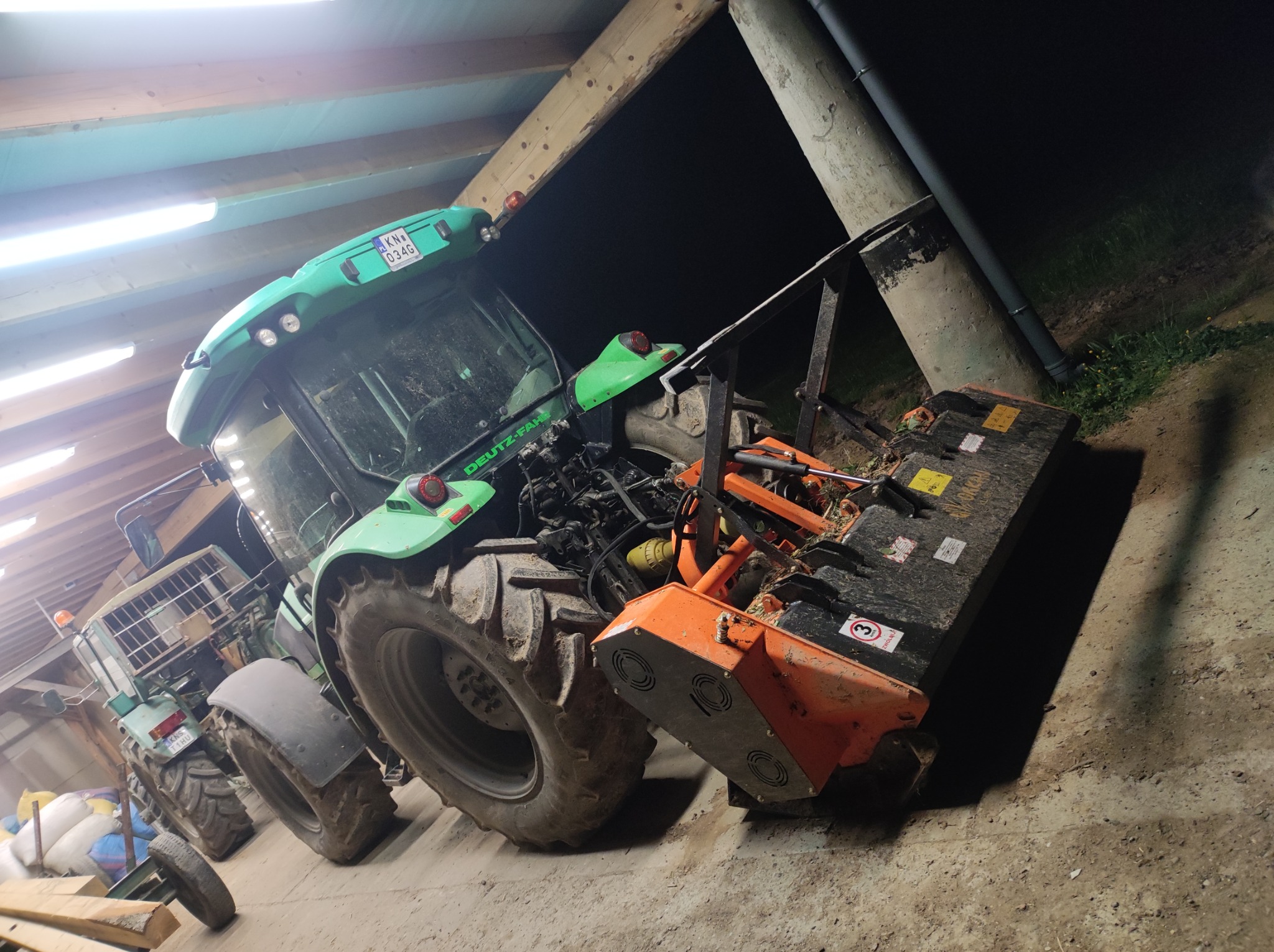 Zielony traktor Deutz-Fahr z pomarańczową kosiarką bijakową wewnątrz budynku, widoczne oświetlenie sufitowe i betonowy słup wentylacyjny na zewnątrz.
