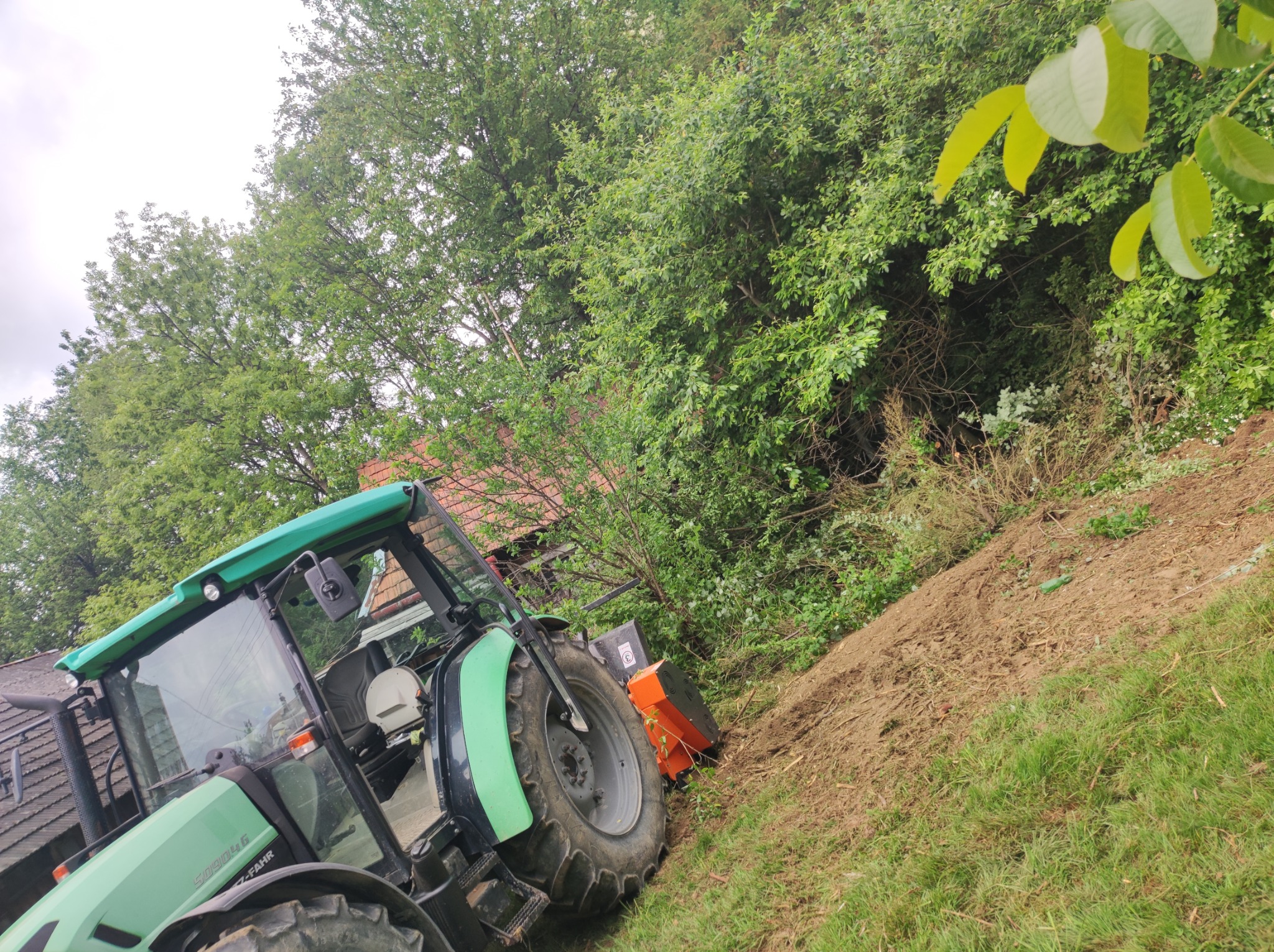 Zielony traktor z pomarańczowym mulczerem na zboczu porośniętym drzewami i krzewami, w tle widoczny dach budynku.
