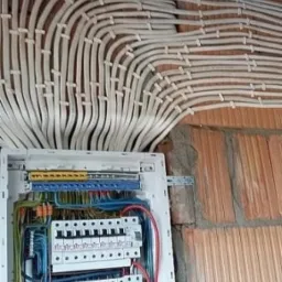 Rozdzielnica elektryczna z licznymi przewodami ułożonymi równolegle na ścianie z cegły.