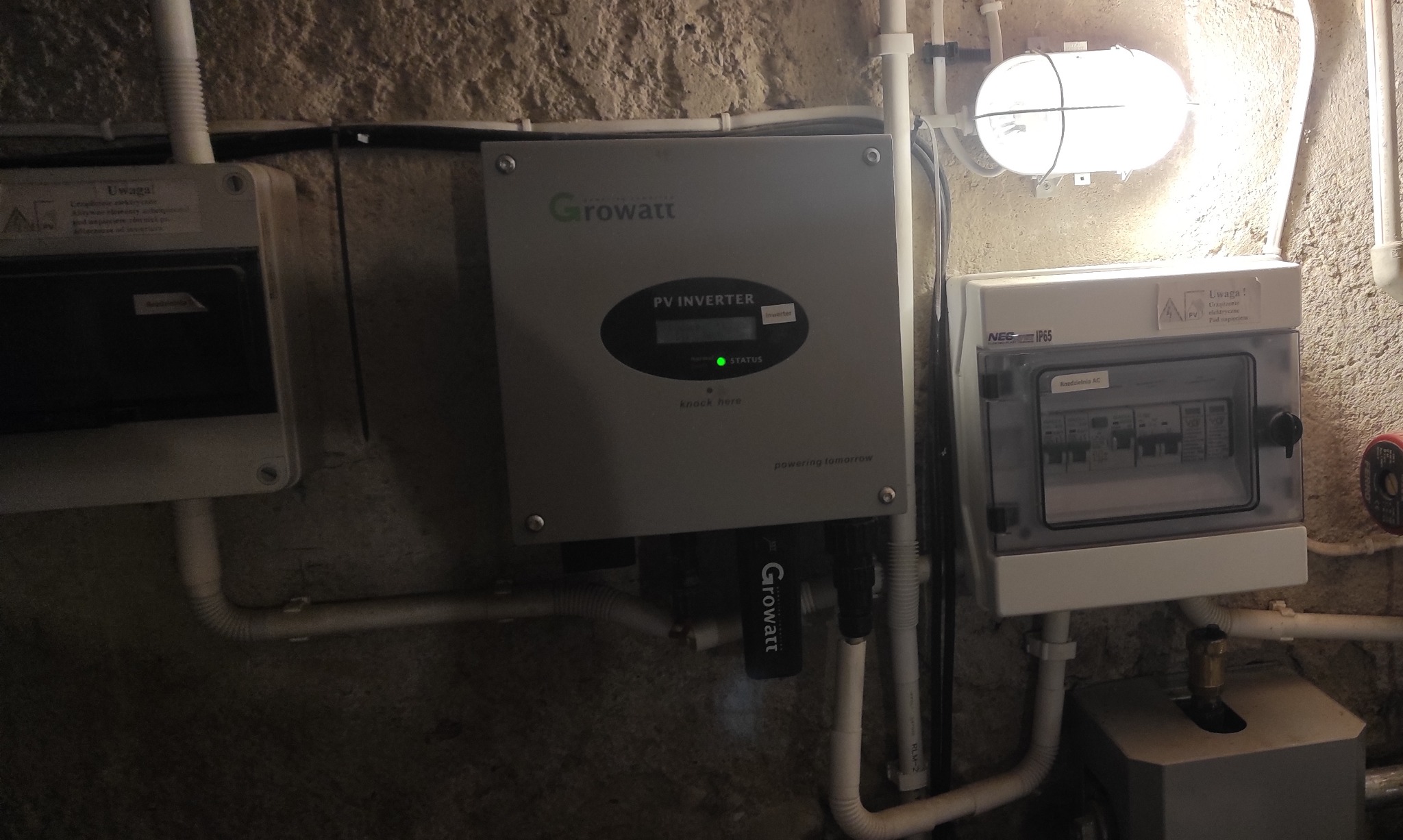 Szary falownik Growatt PV Inverter z zieloną diodą statusu, zamontowany obok skrzynki rozdzielczej z bezpiecznikami, widoczne rury instalacyjne i oświetlenie w piwnicy w Hajnówce.