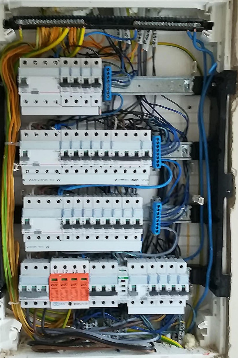 Otwarta skrzynka rozdzielcza z bezpiecznikami, widoczne okablowanie w kolorach żółtym, zielonym, niebieskim i czarnym, oraz listwy zaciskowe.