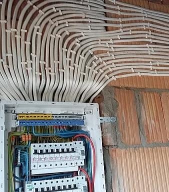 Rozdzielnica elektryczna z licznymi przewodami ułożonymi równolegle na ścianie z cegły.