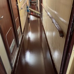 Vanguard Marine - Błyszcząca, brązowa podłoga w wąskim korytarzu z drewnianymi drzwiami i poręczą, w tle widoczne narzędzia i butla z gazem.