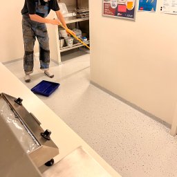 Vanguard Marine - Pracownik w kombinezonie malarskim rozprowadza wałkiem jasną posadzkę z ciemnymi drobnymi plamkami w pomieszczeniu z metalowymi regałami i niebieską kuwetą malarską na podłodze.