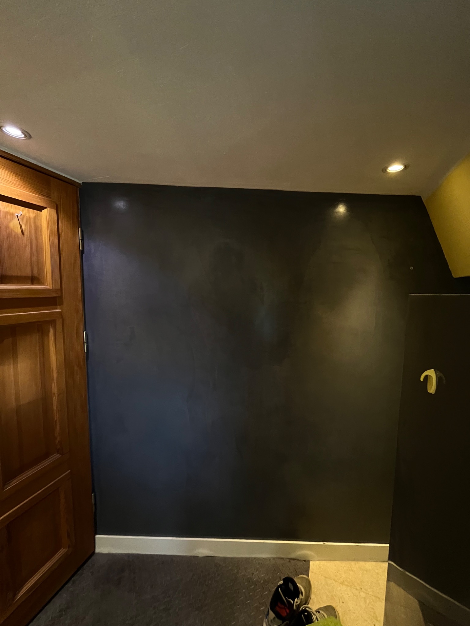 Ciemna ściana z efektem metalicznego połysku, widoczna przy drewnianych drzwiach i fragmentach podłogi z butami.