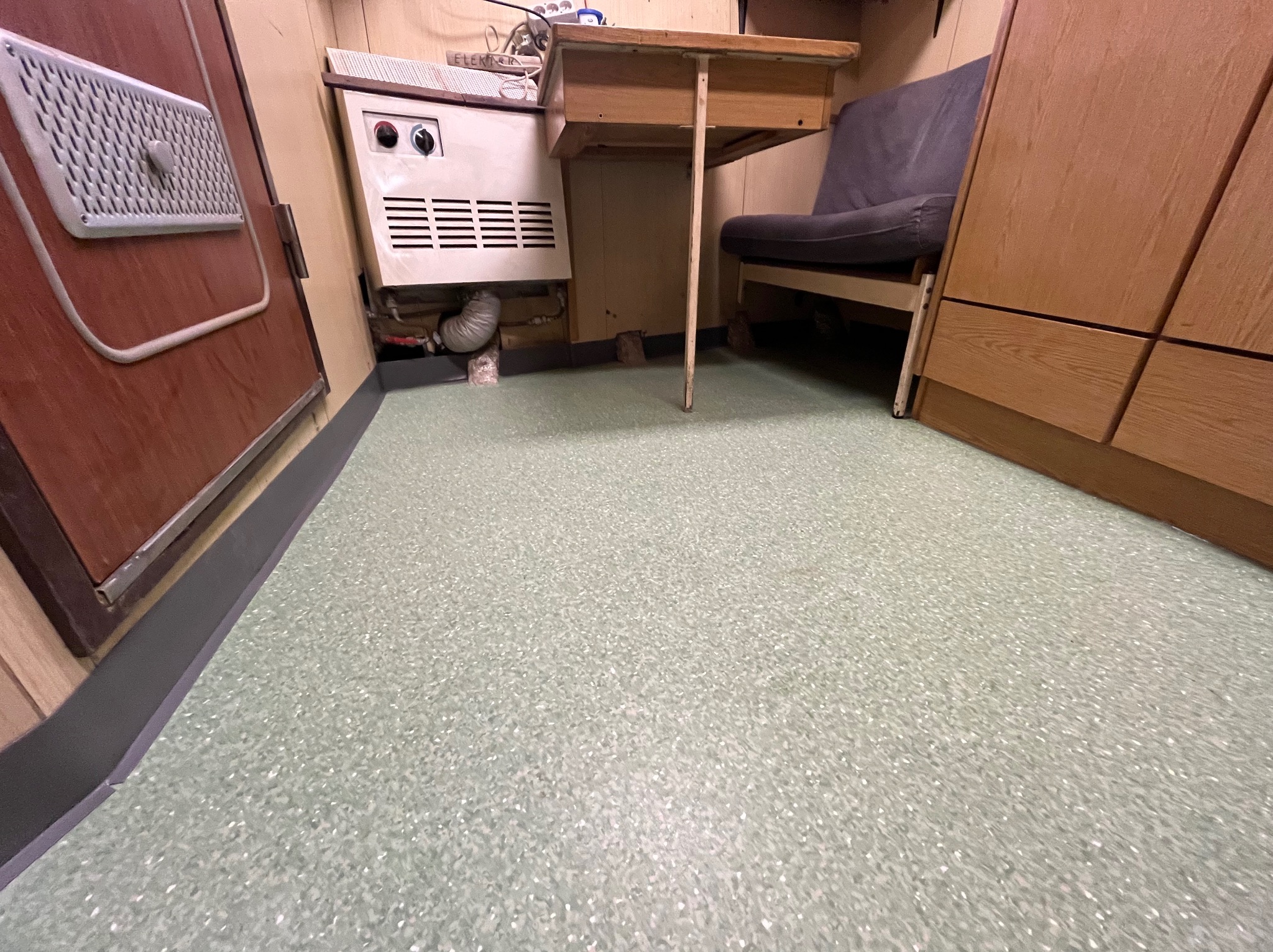 Jasnozielona podłoga z drobnymi plamkami, widoczna przy ścianie z grzejnikiem i starą szafką oraz stolikiem.